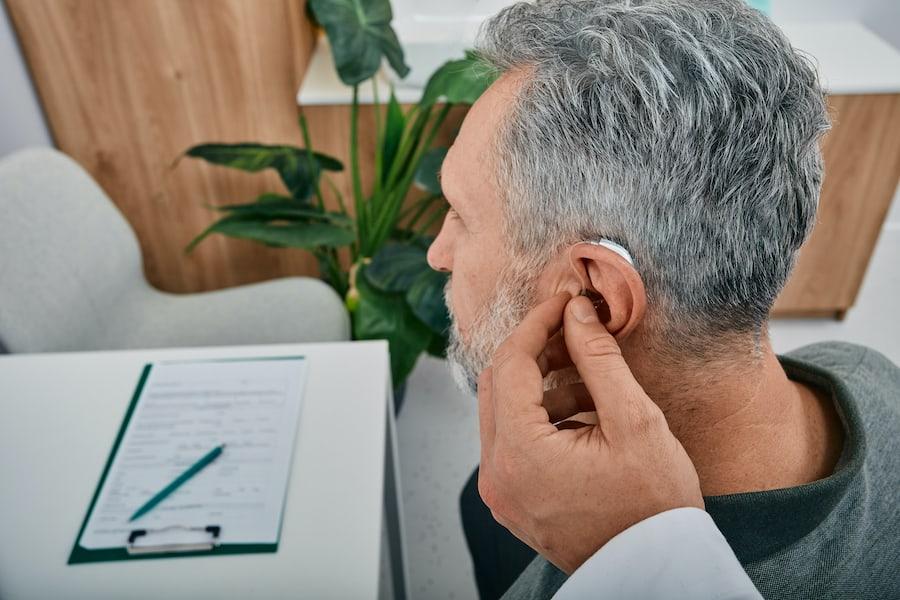 Audiologist Fits A Hearing Aid On Deafness Mature Man Ear While Visit A Hearing Clinic. Hearing Solu